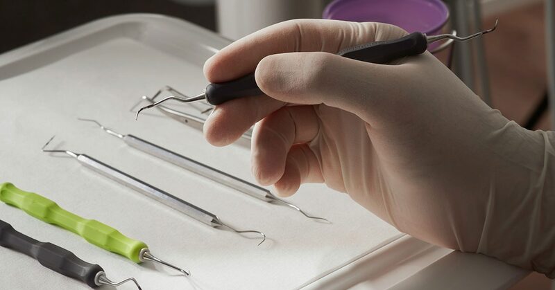 Mano con guante sosteniendo un explorador dental sobre una bandeja con instrumental de revisión